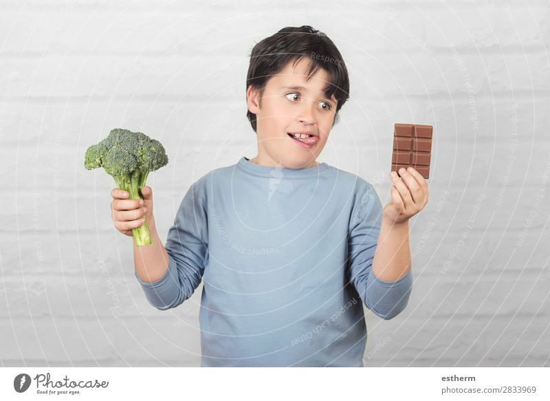 Hungriges Kind mit Brokkoli und einem Schokoriegel in den Händen vor einem Backsteinhintergrund Lebensmittel Gemüse Dessert Schokolade Ernährung Essen