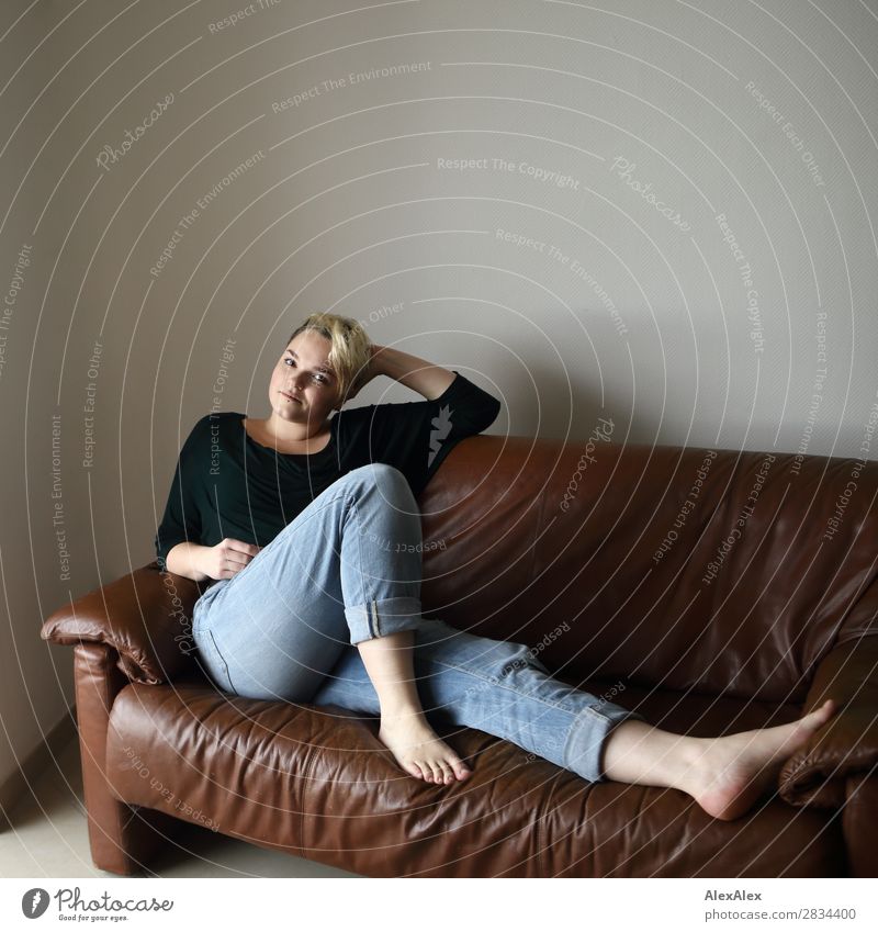 Junge Frau sitzt auf der Couch und streckt ihre langen Beine aus Stil Freude schön Zufriedenheit Sofa Raum Jugendliche 18-30 Jahre Erwachsene Jeanshose Barfuß