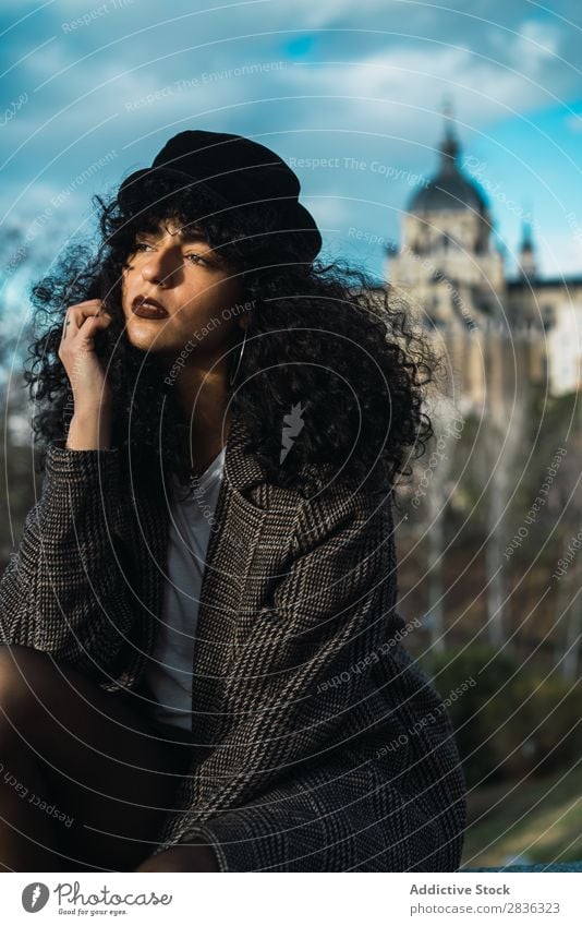 Junge stilvolle Frau, die in der Stadt sitzt. attraktiv Großstadt modisch lockig brünett Mantel Hut sitzen Zaun Aussicht Mode Jugendliche schön hübsch Straße