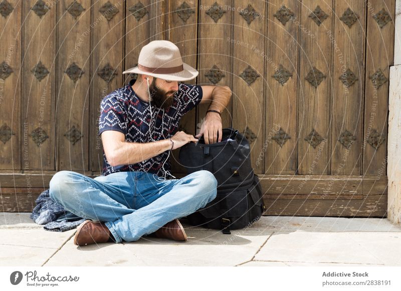 Mann sitzt mit Kopfhörer und Rucksack durchsuchend sitzen Ferien & Urlaub & Reisen Tourismus Boden Porträt Musik Jugendliche Lifestyle Hut bärtig gutaussehend
