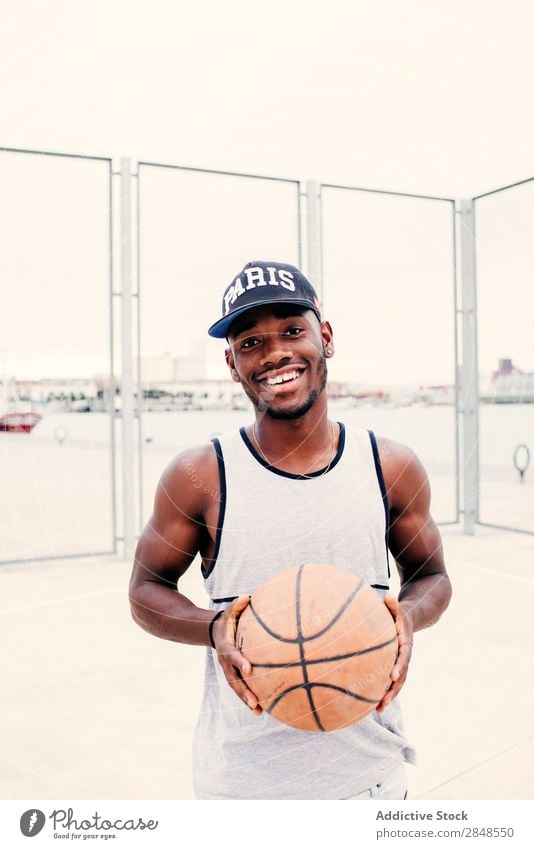 Schwarzer Mann, der lächelnd posiert. Basketball ausdehnend Straßenball selbstbewußt Sportpark Stadt maskulin Sportler Stil Körperhaltung Afroamerikaner