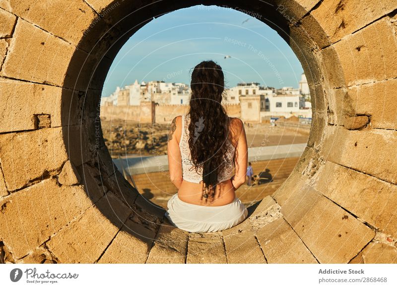 Frau, die auf einem Steinmonument in der Nähe der alten Stadt sitzt. Großstadt Denkmal Marrakesch Marokko antik Felsen Jugendliche sitzen brünett Entwurf Dame