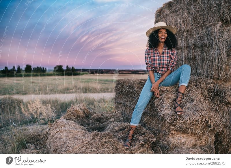 Glückliche schwarze junge Frau, die auf einem Haufen Heu sitzt. Landwirt Sommer urwüchsig Afrikanisch Landschaft Natur Länder Himmel Erholung Lifestyle