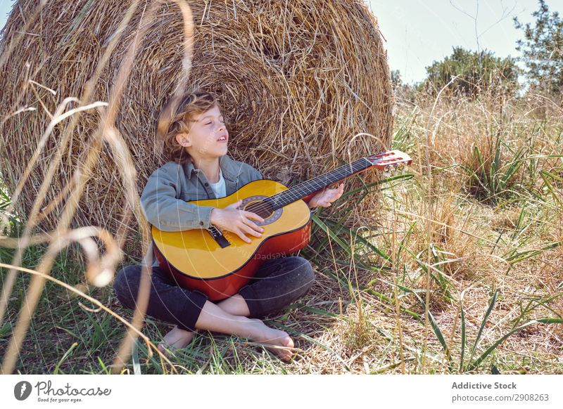 Junge spielt Gitarre und singt kurz vor der Heurolle. Spielen Gesang Musik Feld Brötchen geschlossene Augen Landschaft Kind Gras regenarm Lied akustisch