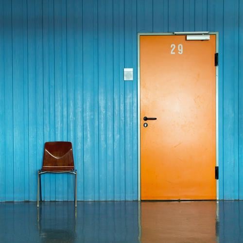 orangene Tür mit Nummer 29 und Türschild in blauer Wand und einem Stuhl für Wartende  davor. Büro Klassenraum Flur Eingang Ziffern & Zahlen warten ästhetisch