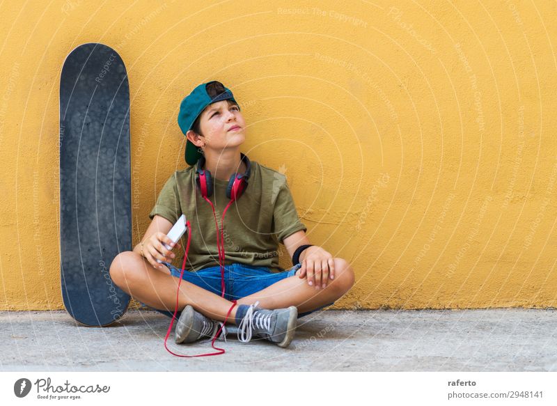 Vorderansicht eines Jungen, der auf dem Boden sitzt und sich an eine gelbe Wand lehnt, während er ein Mobiltelefon benutzt und Musik hört Kind Telefon PDA