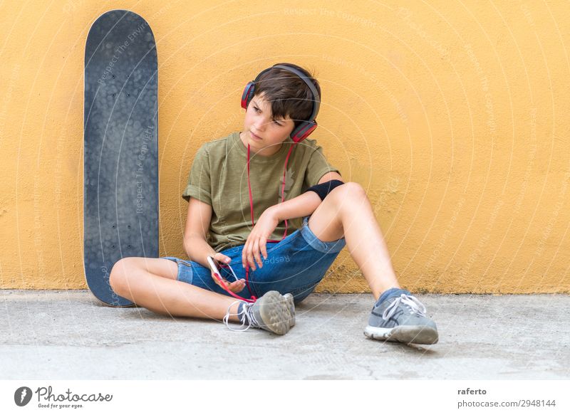 Vorderansicht eines jungen Skaterjungen, der an einer gelben Wand sitzt und über Kopfhörer Musik hört Lifestyle Stil Glück schön Freizeit & Hobby Freiheit