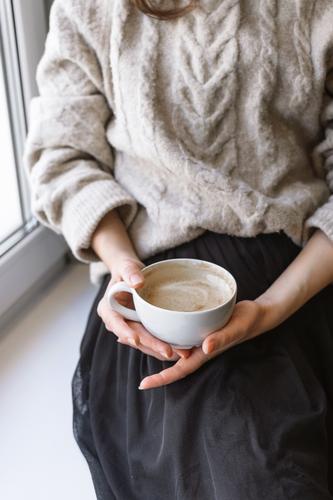 Damenhaltebecher mit Latte vor dem Fenster Herbst Winter Erholung Pullover Frühstück Innenaufnahme Hand Tasse Becher Wärme gemütlich Café Nahaufnahme Kaffee