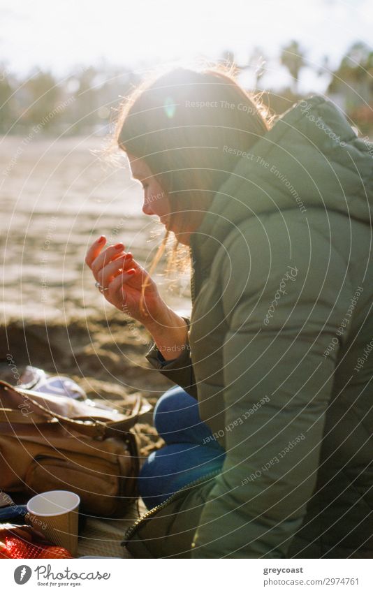 Eine junge Frau in einem warmen Mantel, die auf einem Boden in einem Park sitzt und einen kleinen Snack zu sich nimmt Freizeit & Hobby Mensch Junge Frau