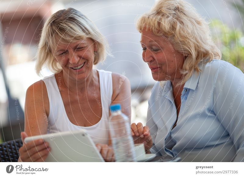 Zwei Frauen mittleren Alters sitzen in einem Straßencafé und lächeln zu etwas in ihrem Block Trinkwasser Limonade Flasche Freude Computerspiel sprechen Mensch