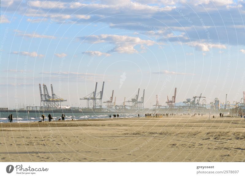 Menschen spazieren am Strand von Valencia im Winter, Spanien. Szene mit Industriehafen und Containerkränen Meer Himmel Wellen Küste Binnenschifffahrt Hafen