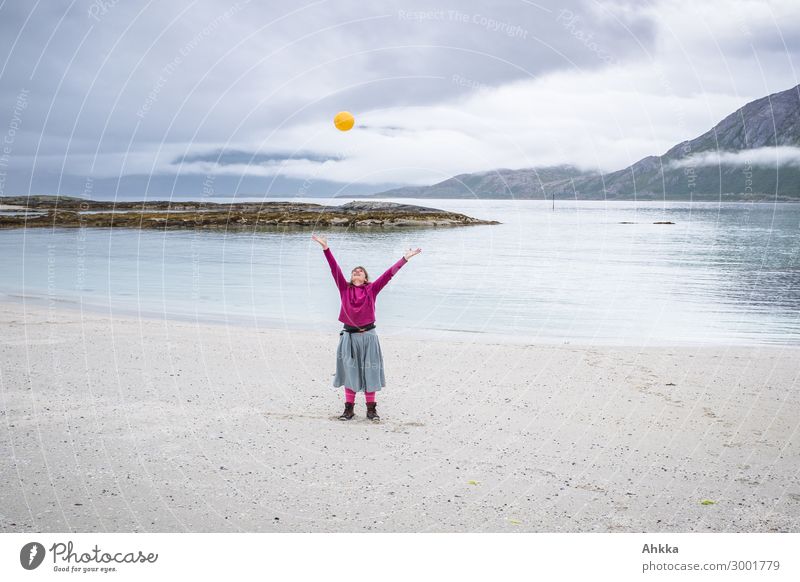 Junge Frau mit ausgebreiteten Armen wirft einen gelben Ball in die Luft am Strand an einem Fjord Konzentration sorgenfrei Unbeschwertheit werfen Freiheit Regen