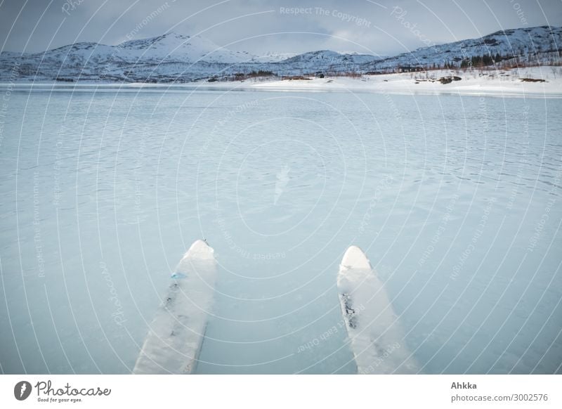 Zwei Skispitzen die mittig von unten ins Bild ragen und unter Wasser eines Sees liegen, die Spitzen schauen aus dem Wasser, vereister See, Blautöne, verschneite Berglandschaft