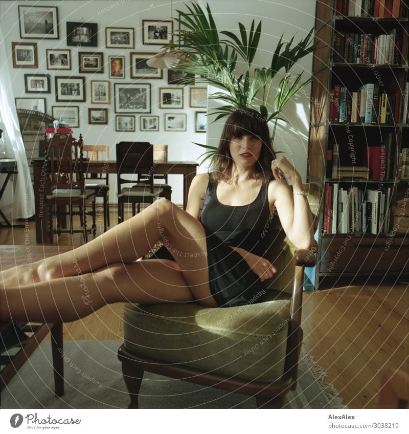 Junge Frau im Wohnzimmer auf einem Sessel Lifestyle elegant Stil schön Erholung Häusliches Leben Möbel Bücherregal Bilderrahmen Zimmerpflanze Jugendliche Beine