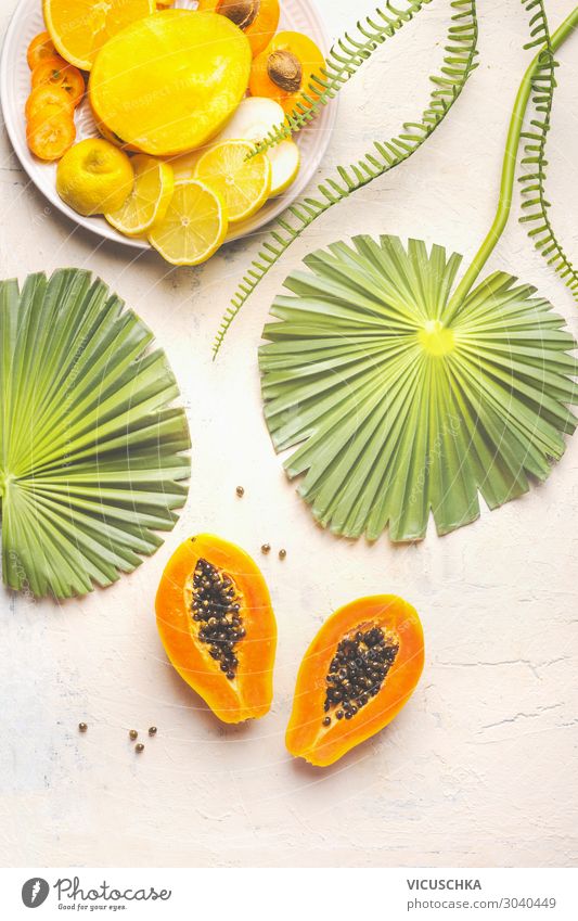 Halbierte reife Papayafrucht mit Samen auf weißem Tisch mit tropischen Blättern und Teller mit gelben geschnittenen Früchten, Draufsicht. Sommerliches Essen. Gesunde Ernährung. Frühstücks-Früchteteller. Modern. Raum kopieren