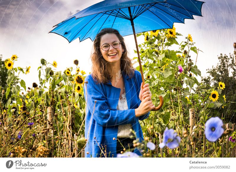 Lächelnde junge Frau mit blauen Regenschirm und Sonnenblumen im Hintergrund feminin Junge Frau Jugendliche 1 Mensch Pflanze Wassertropfen Himmel Wolken Sommer