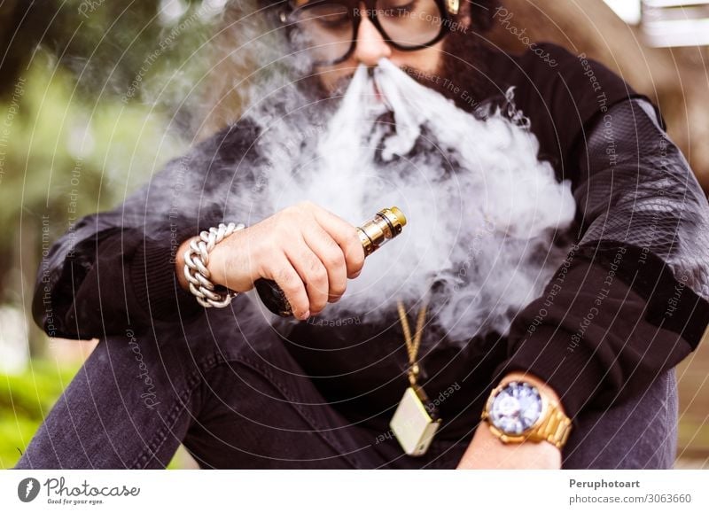 Mann mit Bart raucht eine elektronische Zigarette. Lifestyle Mensch Erwachsene Hand Natur Park Mode Bekleidung Sonnenbrille Hut Vollbart sitzen Coolness modern