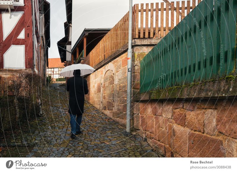 Mann mit Schirm ruhig maskulin Erwachsene 1 Mensch Dorf Fußgänger Wege & Pfade Mode Jeanshose Mantel Regenschirm Mütze Stein Bewegung Erholung gehen historisch