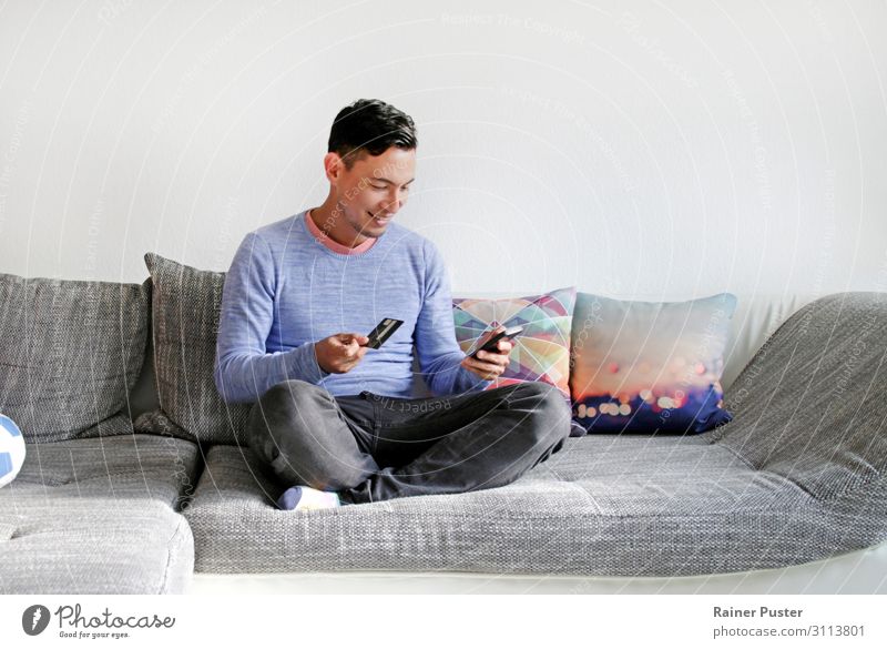 Dunkelhaariger Mann sitzt auf der Couch mit Kreditkarte und Smartphone in der Hand Wirtschaft Business Mittelstand Onlineshop Telefon Handy PDA