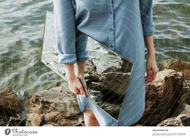 Frau steht auf einem Ufer in blauem Kleid und hält einen Spiegel. Umwelt Sauberkeit Natur Entwurf Erwachsene Felsen Wellen horizontal gesichtslos Küste