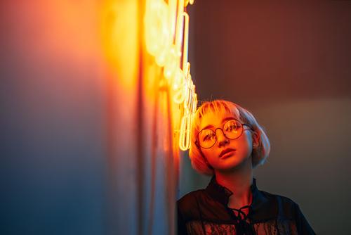 Junge erwachsene Asiatin mit Brille vor einem Neonlichtschild, flacher selektiver Fokus Asiate blond Frau Junge Frau Mädchen Beleuchtung Illumination Licht