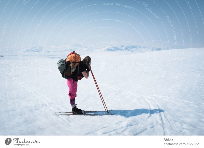 Junge schwer beladene Skifahrerin mit pinker Hose und orangenem Rucksack genießt das verschneite nordische Bergpanorama und ruht sich auf ihren Skistöcken aus