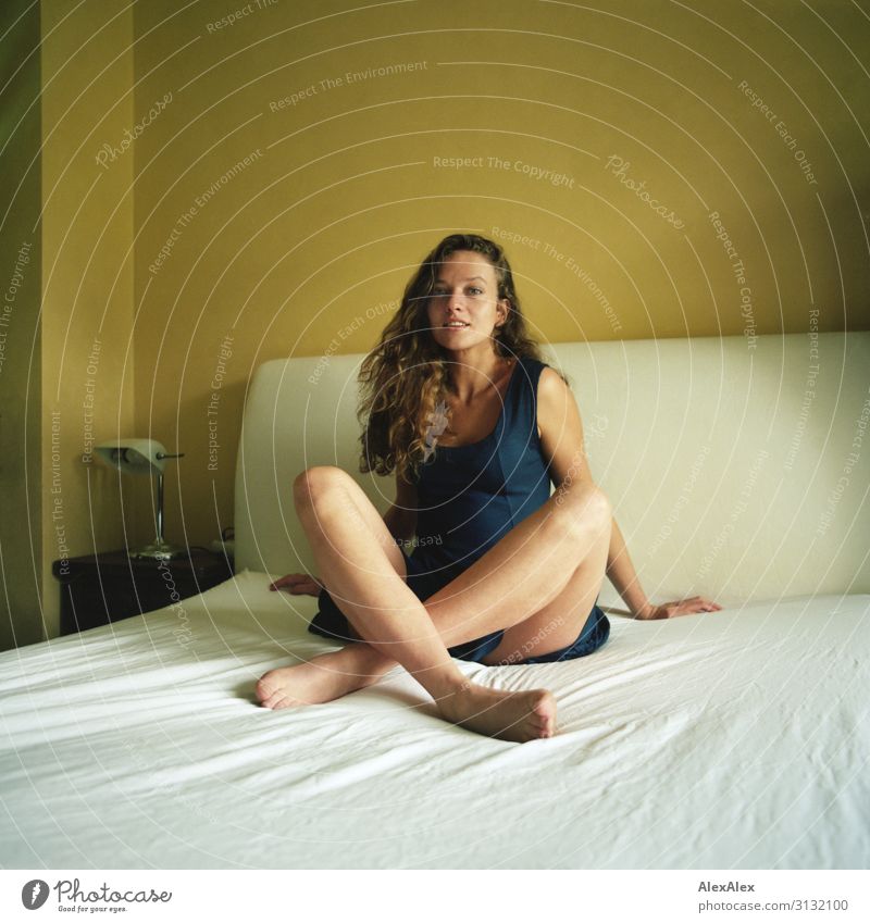 Portrait einer jungen Frau mit langen Beinen auf einem weißen Bett im Schneidersitz Stil schön Leben Wohnung Junge Frau Jugendliche Model 18-30 Jahre Erwachsene