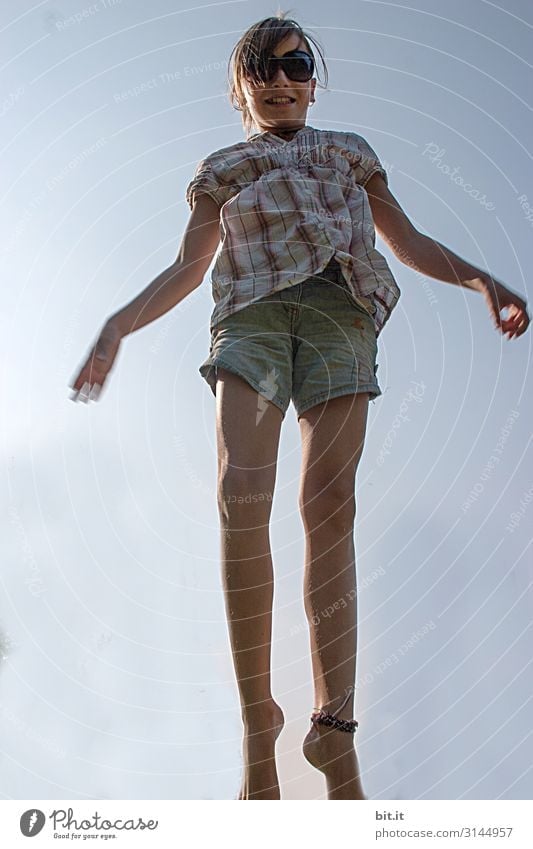 Mädchen mit Sonnenbrille, kurzer Jeans und langen Beinen, hüpft im Sommer fröhlich, glücklich auf dem Trampolin hoch, mit blauem Himmel im Hintergrund.