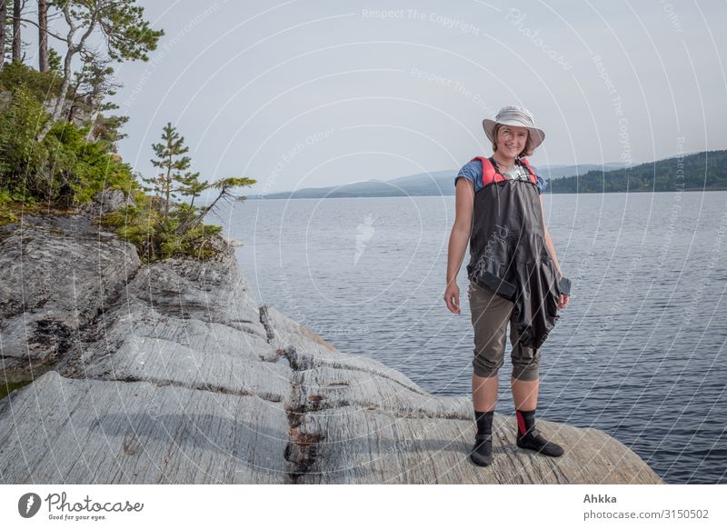 Junge Frau mit Hut steht am Rand eines Felsens an der Uferkante eines Sees und blickt in die Kamera Abenteuer Urlaub Kajakfahrer Natur Wasser
