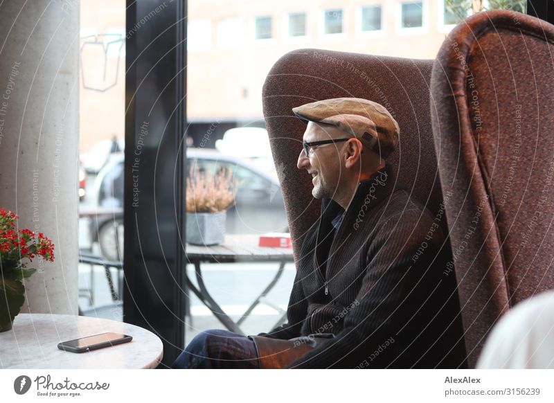 HH UT 19 | Mann sitzt in Ohrensessl im Cafe und lächelt Ernährung Kaffeetrinken Erwachsene 45-60 Jahre Jacke Mütze Brille Tisch Sessel Fenster Telekommunikation