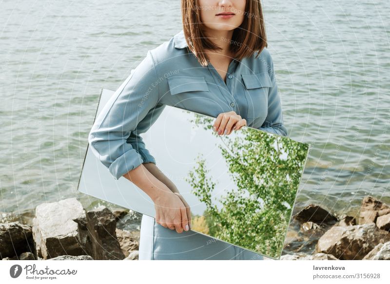 Frau steht auf einem Ufer in blauem Kleid und hält einen Spiegel. Lifestyle Jugendliche 18-30 Jahre Junge Frau Sommer Erwachsene Sauberkeit Entwurf Umwelt