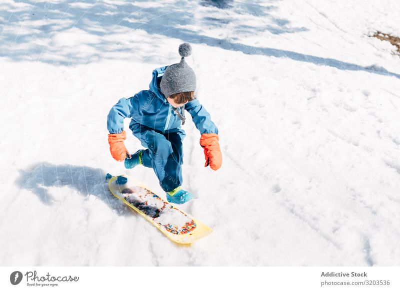 Kind springt an Bord und fährt einen verschneiten Berg hinunter Junge Holzplatte Schnee Rutsche Geschwindigkeit klein Winter Ausritt Sport abwärts Aktion