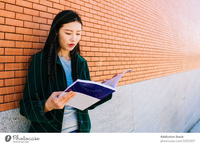 Asiatischer Student liest Lehrbuch in der Nähe der Ziegelmauer Schüler lesen Handbuch anlehnen Wand Backstein asiatisch Frau Studium Jugendliche Notizbuch