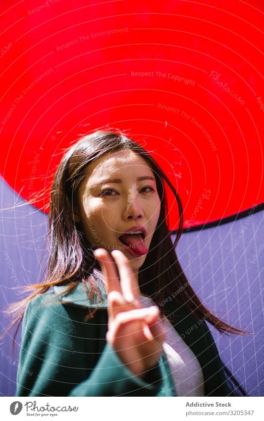 Asiatische Frau mit Zunge und V-Zeichen Zunge zeigen v-Zeichen gestikulieren hell Wand Stadt Großstadt Straße asiatisch Jugendliche lustig zwei Finger Frieden