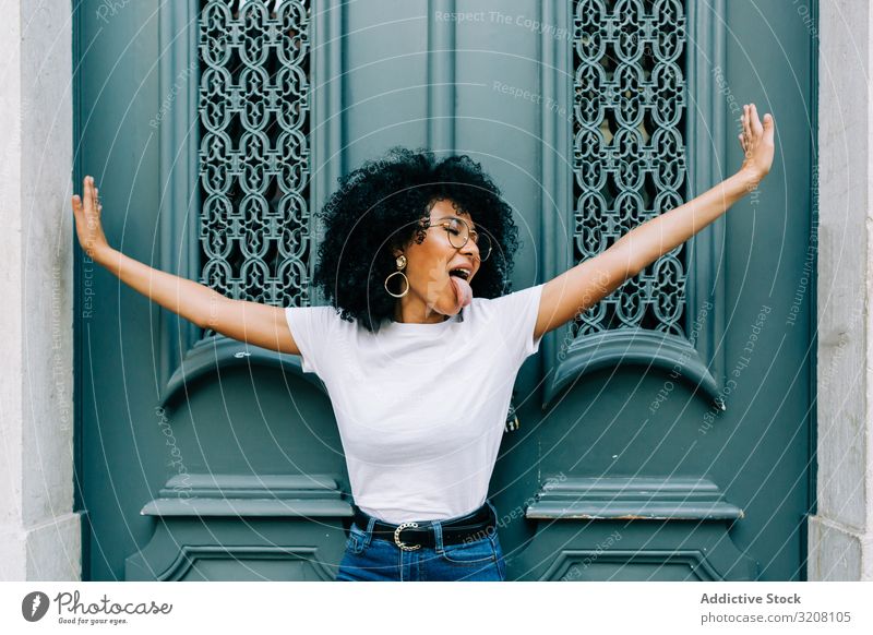 Stilvolle ethnische Frau mit ausgestreckten Armen Tür Zunge klebend spielerisch stylisch trendy Frisur jung Afroamerikaner Person lässig attraktiv schön hübsch
