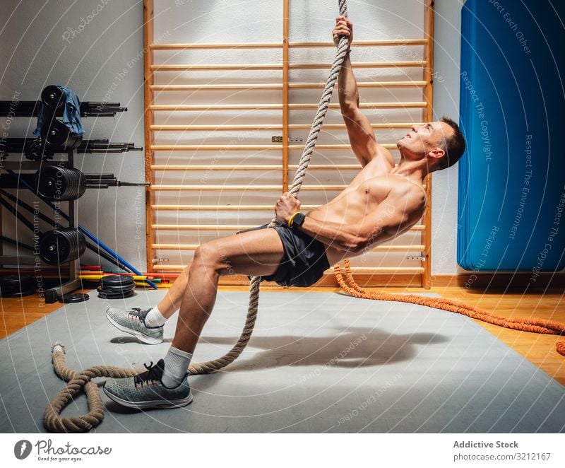 Muskelsportler klettert Seil in Turnhalle Athlet Aufstieg Fitnessstudio Übung Mann Training Sport Stärke Kraft anstrengen physisch modern fokussiert Gerät