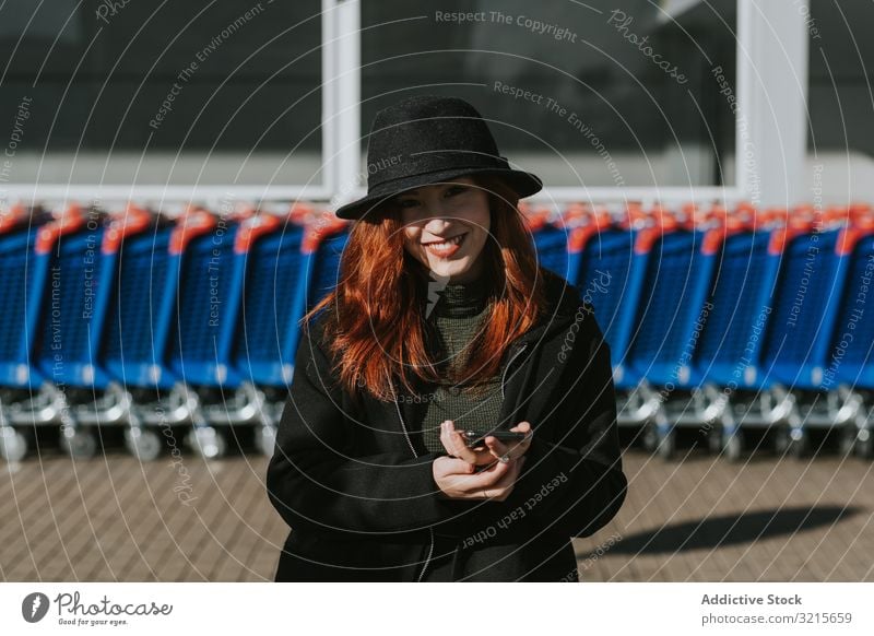 Lächelnde Frau mit Smartphone auf einem Parkplatz mit Einkaufswagen attraktiv jung schön lässig heiter klug Netzwerk Telefon modern Stehen Freude Rotschopf