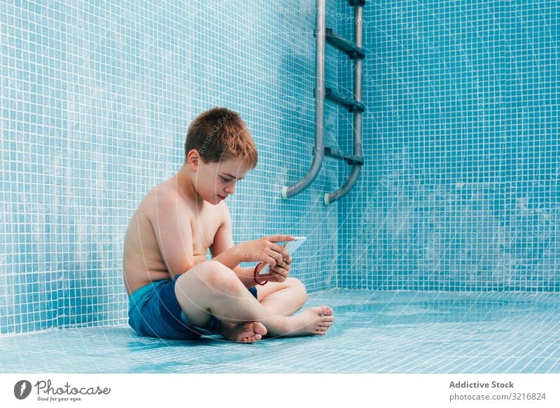 Junge mit Smartphone auf dem Boden eines leeren Schwimmbeckens sitzend Pool Sitzen Gesäß Kind aussruhen Fliesen u. Kacheln dekoriert Technik & Technologie