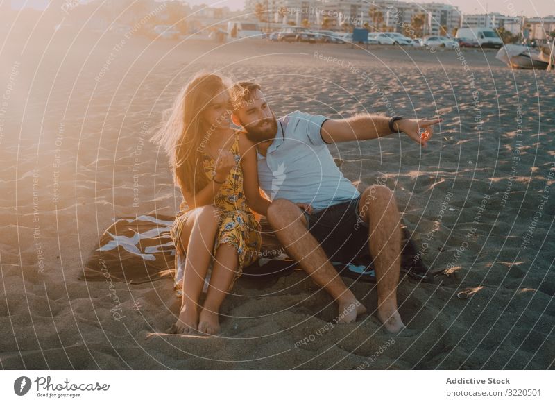 Zärtliches Paar, das sich am Handtuch-Strand im Sonnenlicht unterhält und auf ihn zeigt romantisch Liebe Glück umarmend Angebot Jahrestag Zusammensein Datierung