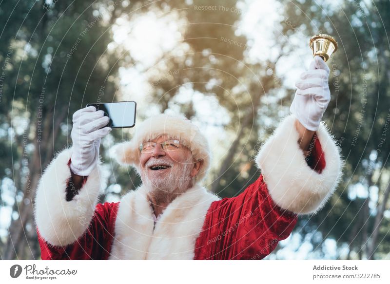 Fröhlicher Weihnachtsmann mit Smartphone und Klingel Selfie Feiertag Mann Weihnachten Veranstaltung benutzend Senior männlich heiter freudig bärtig grauhaarig