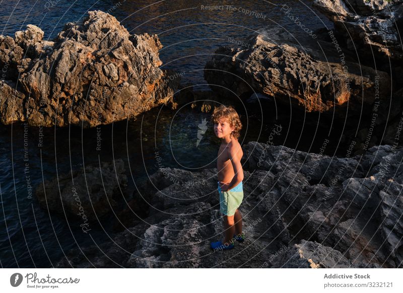 Glücklicher kleiner Junge steht auf einem Felsen am Meeresufer Klippe MEER Lächeln winken Ufer Abenteuer aufgeregt Seeküste sorgenfrei Küste Kind