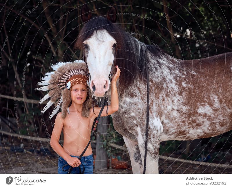 Kind mit authentischem Kopfschmuck umarmt Hengst auf dem Bauernhof Junge Pferd Streicheln Kriegsmütze Art Inder Tracht konzentriert Kraulen ohne Hemd heimatlich