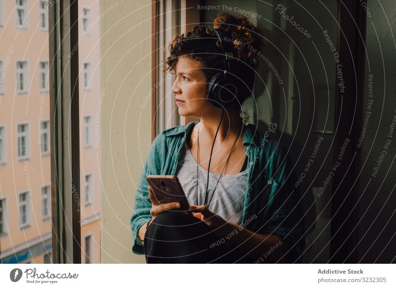 Entspannte Frau mit Kopfhörern und Smartphone zu Hause Lächeln benutzend besinnlich zuhören Glück Musik Browsen Fensterbrett lockig attraktiv friedlich