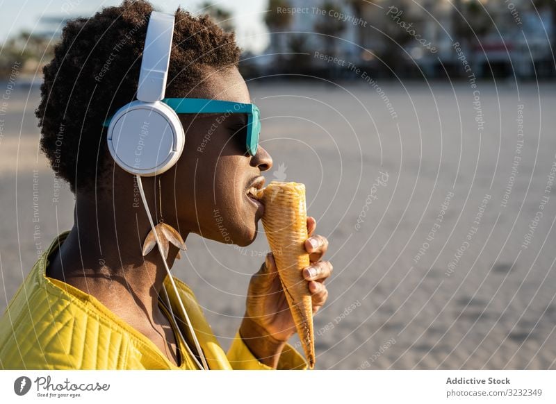Kühle Frau isst Eiscreme am Strand Speiseeis trendy cool essen wüst Afroamerikaner Kopfhörer Jacke gelb hell Sonnenbrille genießen süß Lebensmittel Sommer Spaß