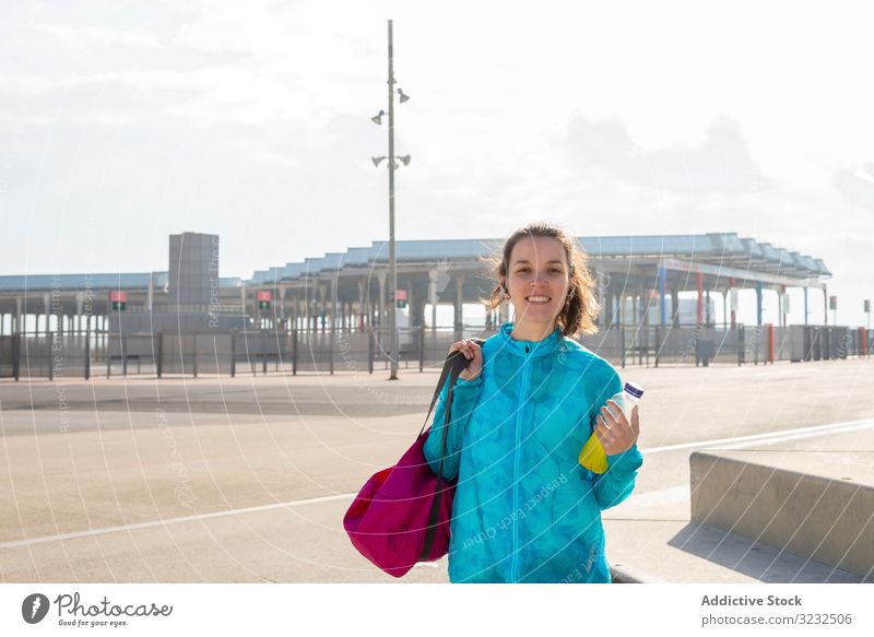 Lächelnde Sportlerin hält Tasche und Flasche, während sie im Freien steht Frau Straße laufen Großstadt Freizeit Stil urban Aktivität Athlet sportlich Körper