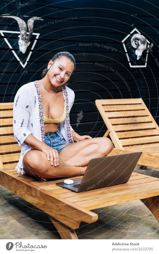 Entspannte Frau benutzt Laptop, während sie sich auf Liegestuhl ausruht ruhen entspannt benutzend Browsen heiter Lächeln hölzern Terrasse tropisch sitzen lässig