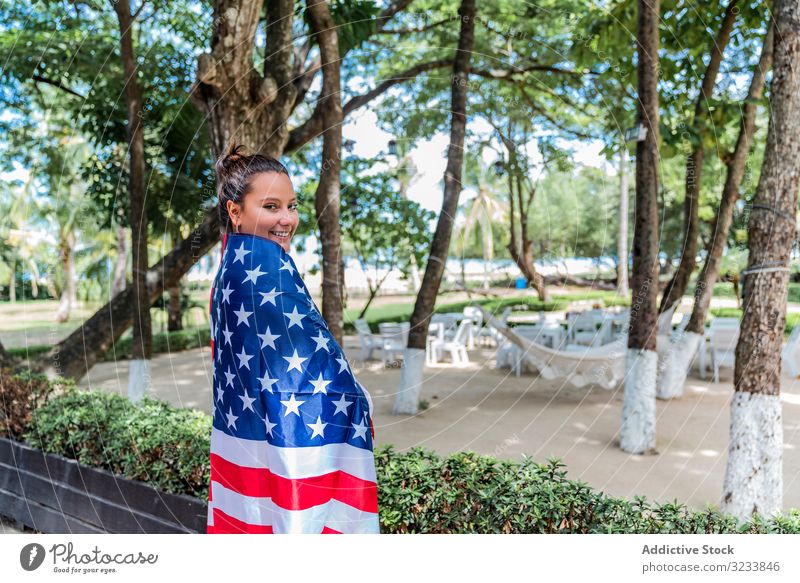 Zufriedene Frau lächelt eingehüllt in die amerikanische Flagge und geht am Park entlang Stars and Stripes national Inhalt Lächeln heiter führen eingewickelt Kap