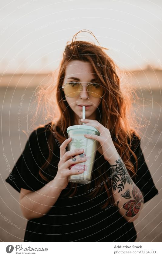 Langhaarige Teenagerin trinkt Milchshake und schaut in die Kamera Frau tausendjährig heiter cool Stil trinken Glück Sonnenuntergang Tattoo Sonnenbrille Piercing