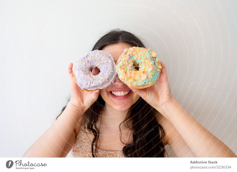 Verspielte Frau macht Gläser mit Donuts, während sie an der weißen Wand steht Krapfen spielerisch Brille Spaß haben sorgenfrei spielen Gebäck verglast Streusel