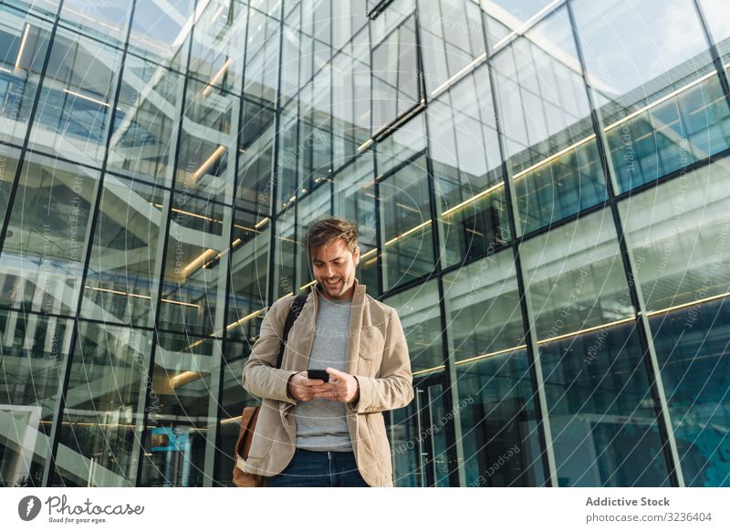Lächelnder Unternehmer nutzt Smartphone vor Bürogebäude Geschäftsmann Straße Großstadt benutzend positiv modern Erwachsener Business Zentrum Hand-in-Tasche